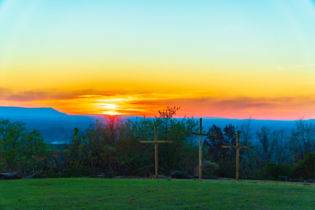 The mountain-top ceremony site at Peaceful Pointe, featuring three rustic wooden crosses with a vibrant sunset and panoramic views of the Arkansas landscape.