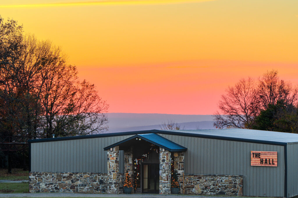 The exterior of The Hall at Peaceful Pointe wedding venue, framed by a vibrant sunset and trees, showcasing a rustic stone entrance and festive lighting.